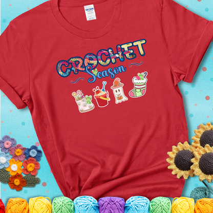 Red t-shirt shows the words “Crochet Season” with cute seasonal icons below: a spring boot with hooks, summer bucket with crab, fall ghost with pumpkin hat, winter stocking, and tiny frogs.