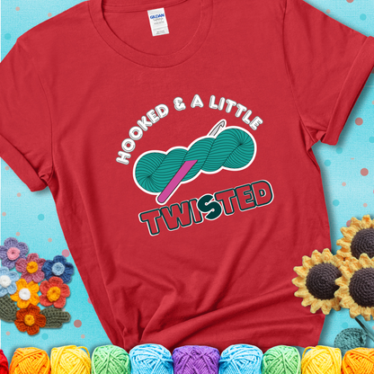 A teal skein of yarn with a hot pink crochet hook through it, captioned “Hooked & a Little Twisted” on a red t-shirt.