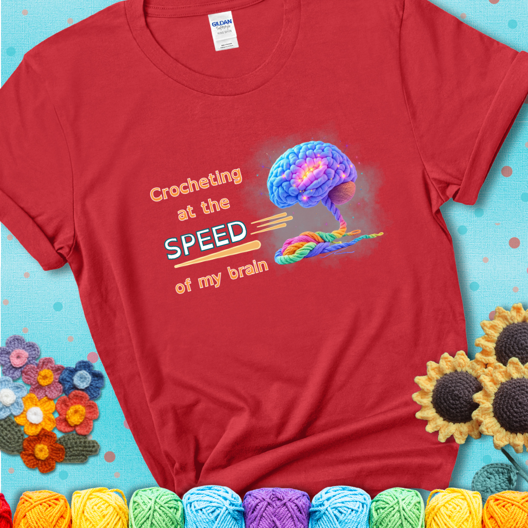 A glowing rainbow brain unravels into colorful yarn with the phrase “Crocheting at the speed of my brain” on a red t-shirt. 