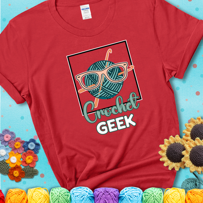 A teal yarn ball with crisscrossed hooks wears oversized nerdy glasses with the phrase “Crochet Geek” in bold, fun text below on a red t-shirt.
