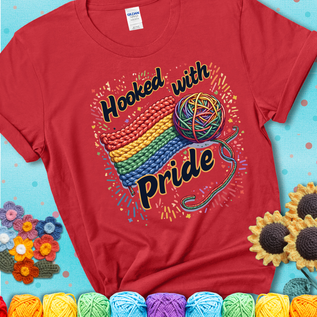 A rainbow-striped crochet swatch paired with a multicolor yarn ball and bold “Hooked With Pride” lettering. Confetti surrounds the vibrant scene showing a red tee with a teal sunflower background