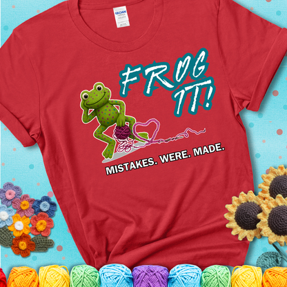 Green frog with yarn and heart-shaped loop. Text reads: “Frog It! Mistakes Were Made.” A playful crochet tee for anyone who's ever had to rip it back, showing a red tee with a teal sunflower background