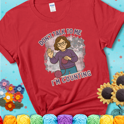 Red t-shirt with an annoyed maker holding up a hand beside yarn, with the phrase “Don’t Talk To Me, I’m Counting.”