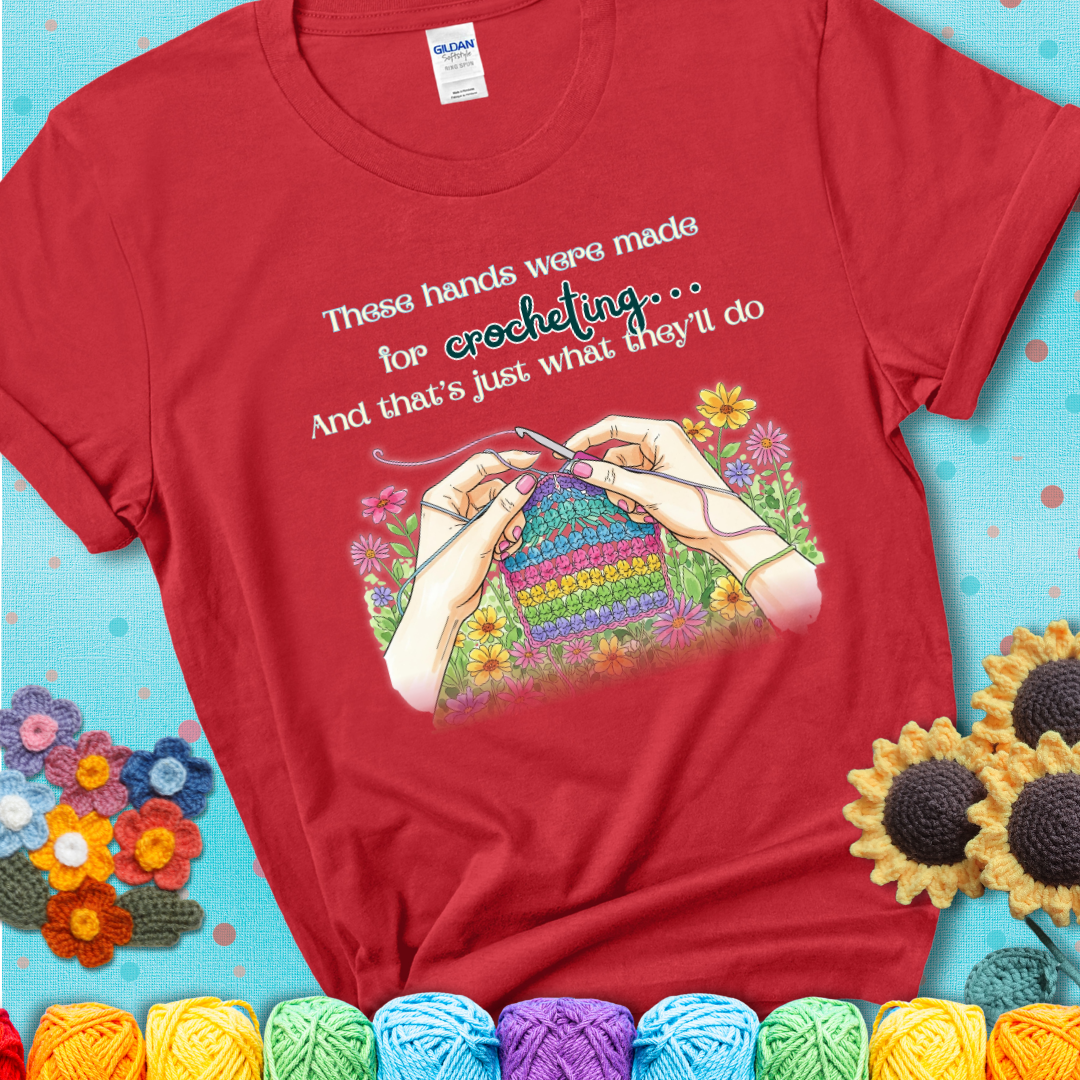 Red t-shirt with an illustration of two hands crocheting a rainbow swatch, surrounded by colorful flowers and the phrase “These hands were made for crocheting… and that’s just what they’ll do.”