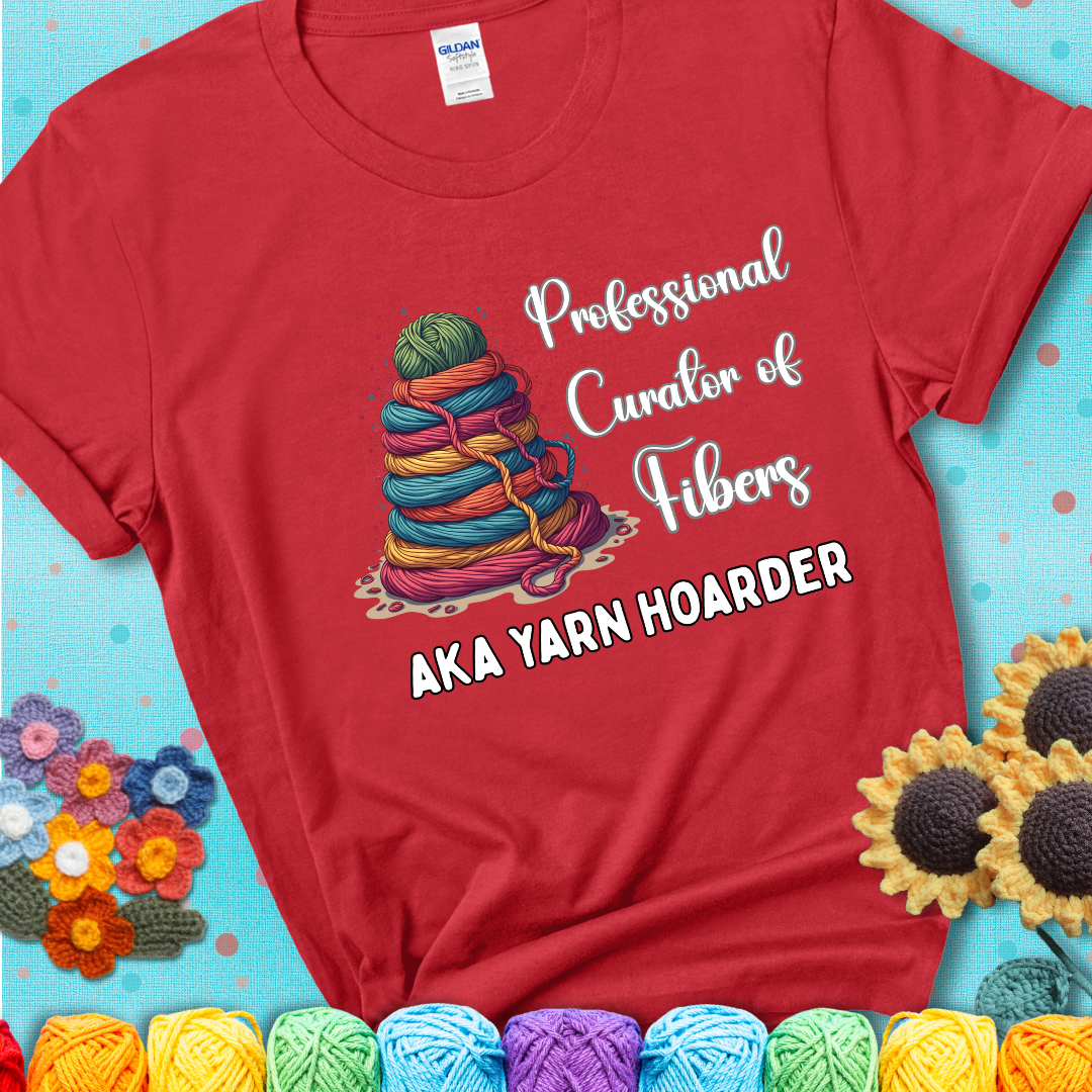 A red t-shirt showing a pyramid of rainbow yarn topped with green ball; text reads “Professional Curator of Fibers AKA Yarn Hoarder”, a playful shirt for serious stashers.