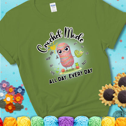 Green t-shirt with a kawaii-style rainbow yarn character surrounded by crochet stitch abbreviations and holding a yarn heart, with text “Crochet Mode All Day, Every Day.”