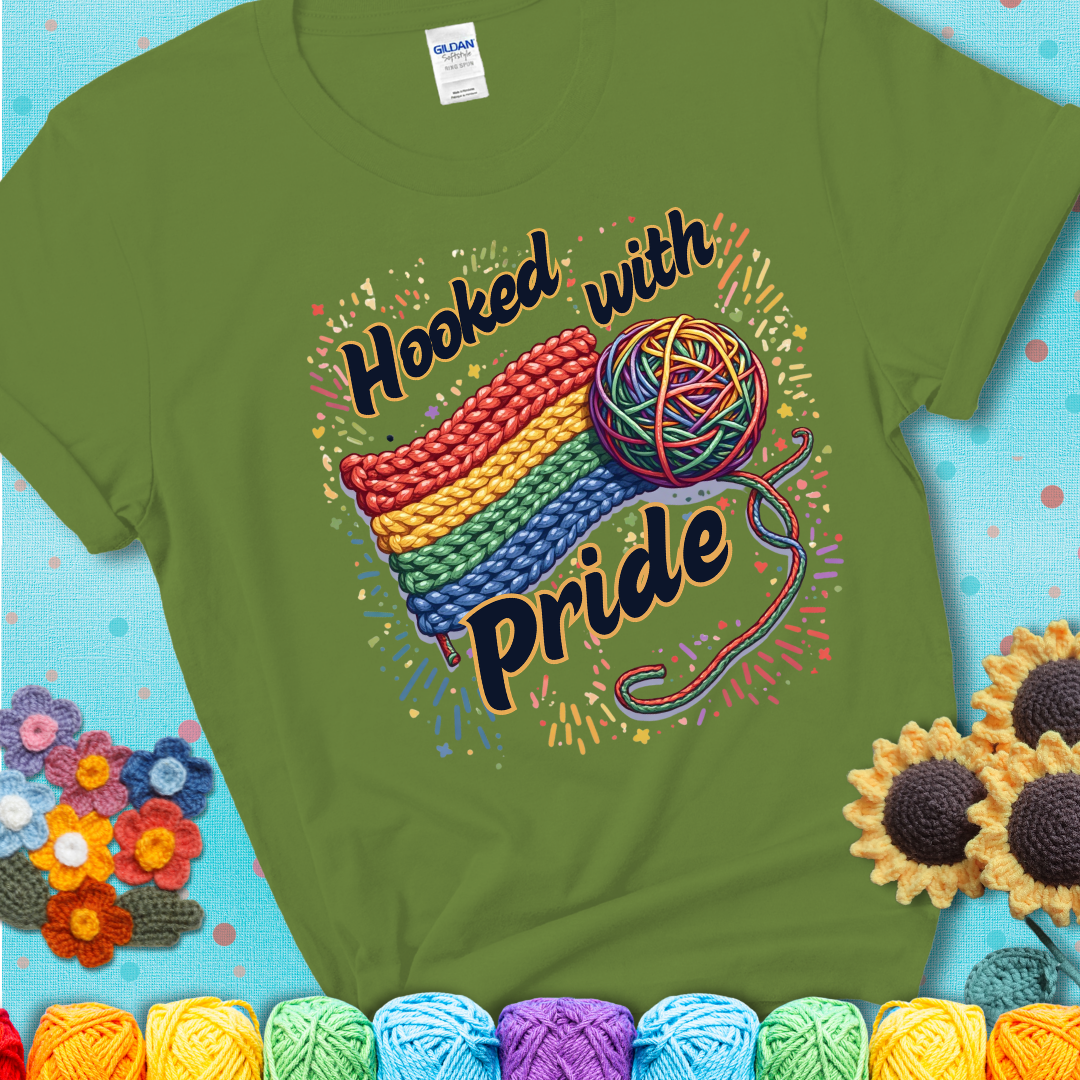 A rainbow-striped crochet swatch paired with a multicolor yarn ball and bold “Hooked With Pride” lettering. Confetti surrounds the vibrant scene showing a green tee with a teal sunflower background