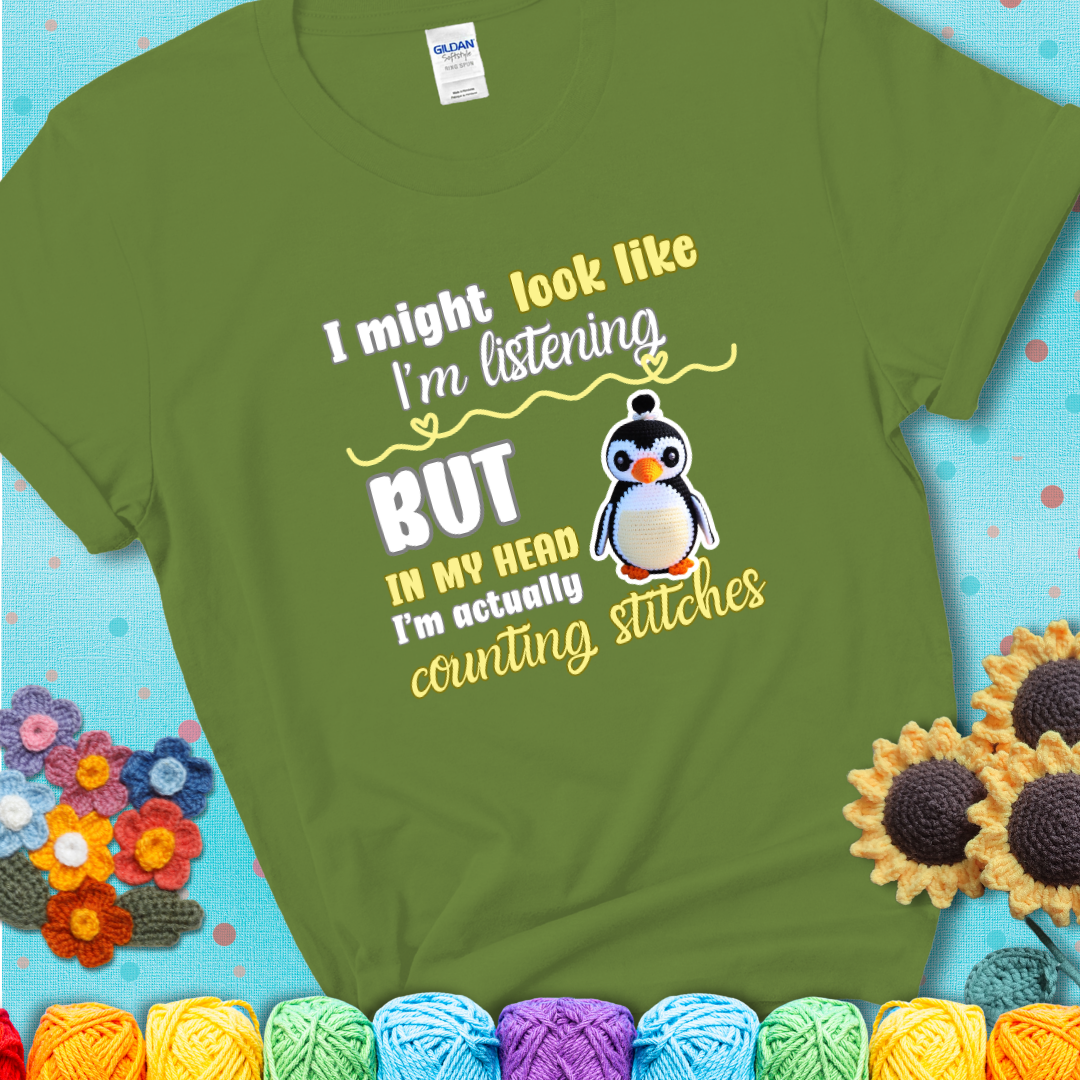 Green t-shirt shows a crocheted penguin next to the text “I might look like I’m listening but in my head I’m actually counting stitches.”