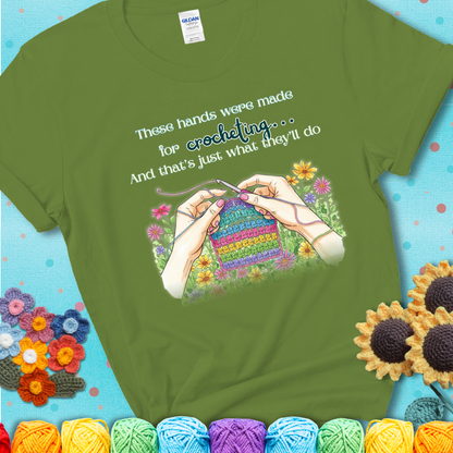 Kiwi t-shirt with an illustration of two hands crocheting a rainbow swatch, surrounded by colorful flowers and the phrase “These hands were made for crocheting… and that’s just what they’ll do.”