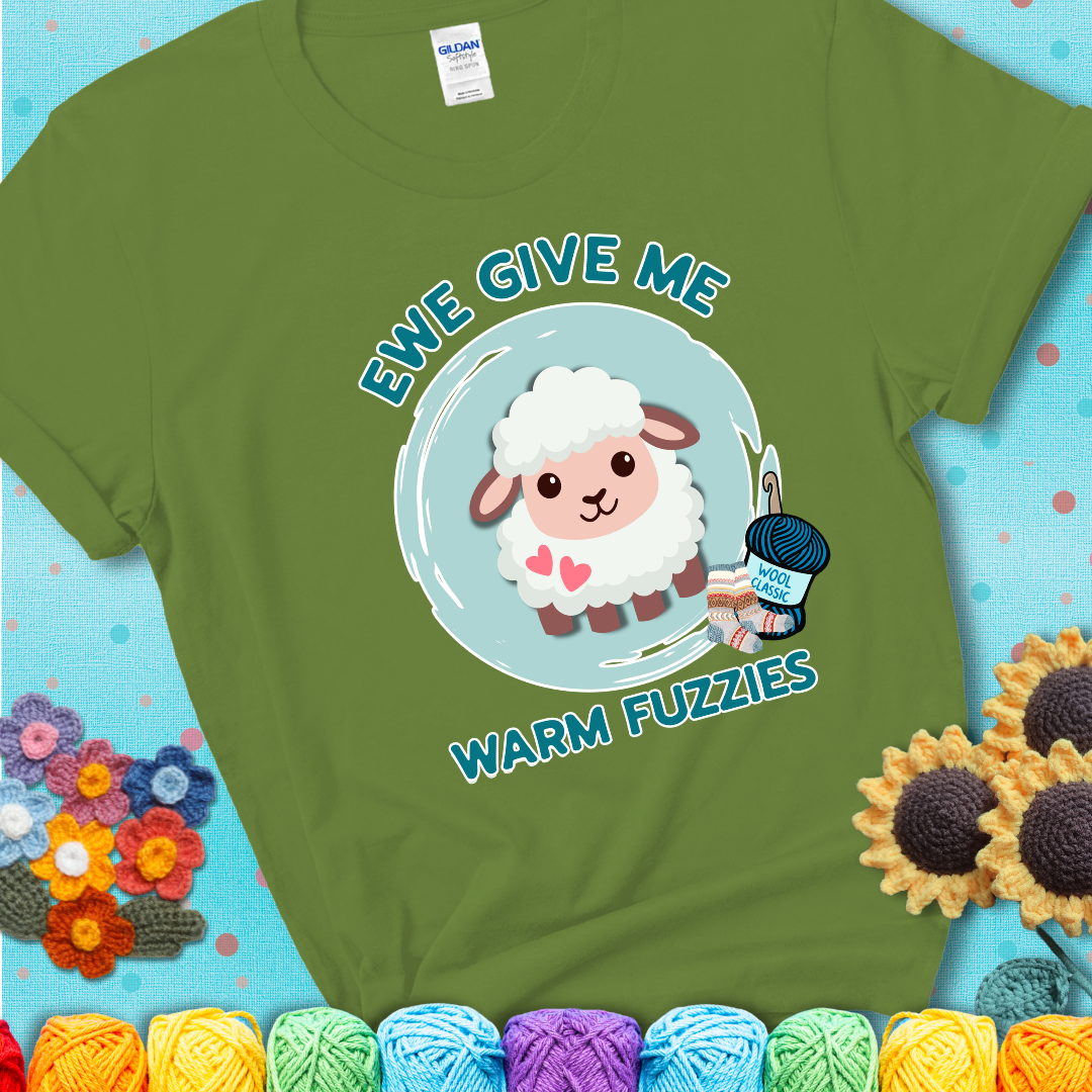 Green t-shirt showing a cartoon sheep with pink hearts, wool socks, and yarn labeled “Wool Classic.” Text reads “Ewe Give Me Warm Fuzzies.”