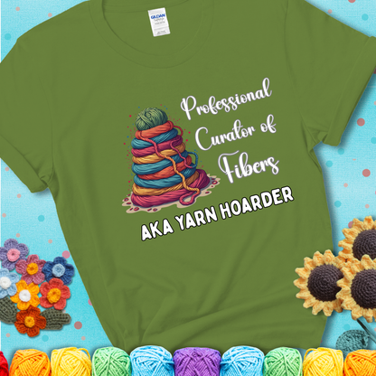A green t-shirt showing a pyramid of rainbow yarn topped with green ball; text reads “Professional Curator of Fibers AKA Yarn Hoarder”, a playful shirt for serious stashers.
