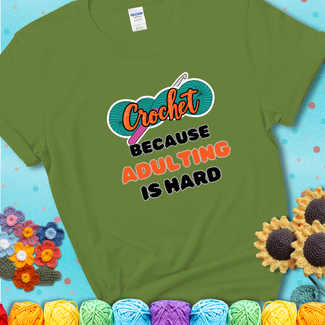 Green t-shirt featuring a bold design with the phrase “Crochet Because Adulting Is Hard” featuring a green skein of yarn with a pink crochet hook. The word “Crochet” is in a script font, while “Adulting is hard” is in chunky textured letters.