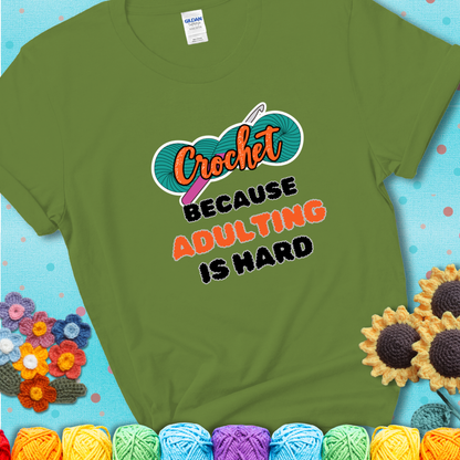 Green t-shirt featuring a bold design with the phrase “Crochet Because Adulting Is Hard” featuring a green skein of yarn with a pink crochet hook. The word “Crochet” is in a script font, while “Adulting is hard” is in chunky textured letters.