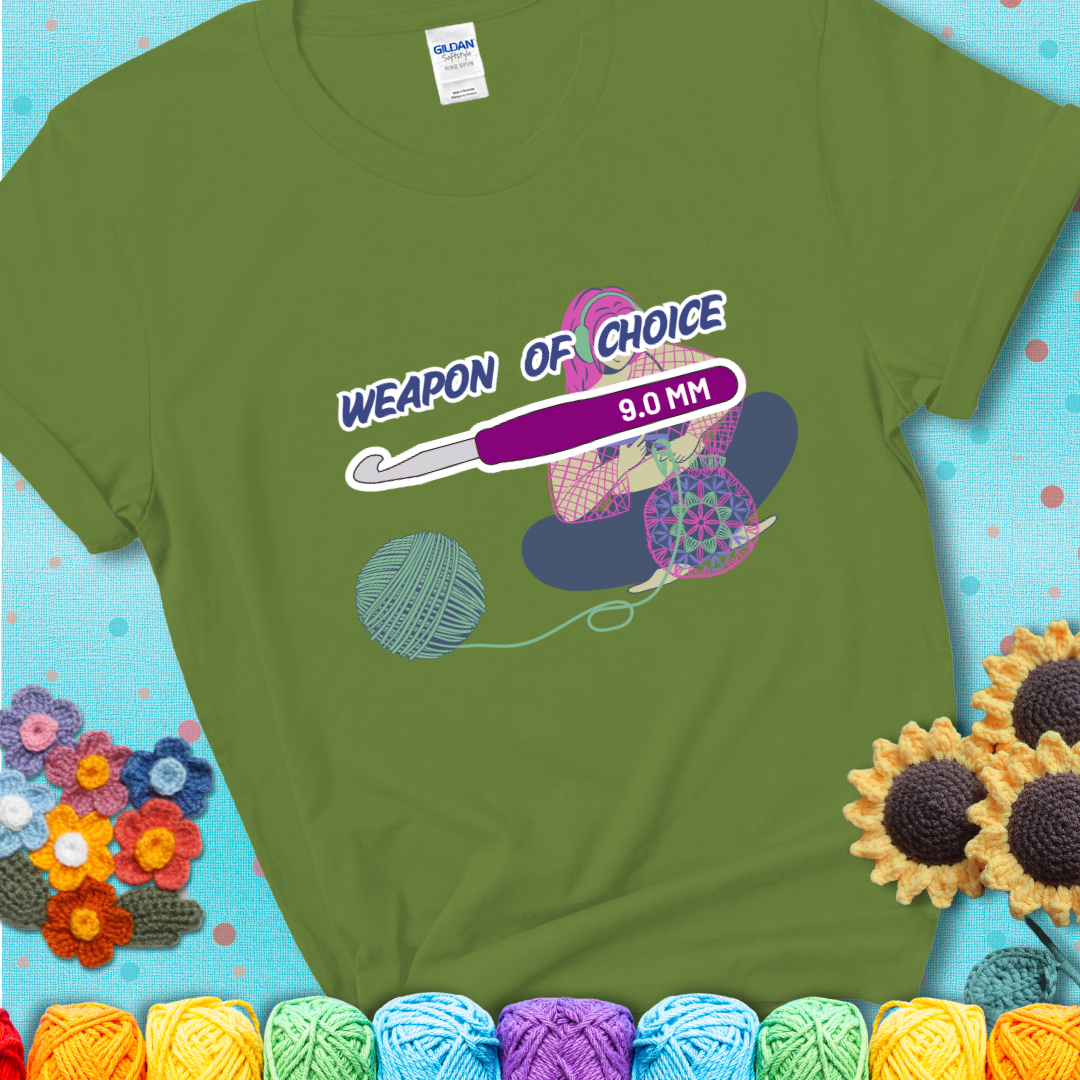 Green t-shirt with an Illustration of a crocheter sitting cross-legged with a 9.0 mm crochet hook above her, labeled “Weapon of Choice.” She’s crocheting a colorful mandala-style project from a large yarn ball.