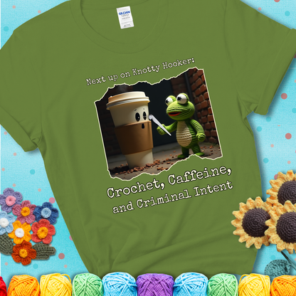 A crocheted frog holding a crochet hook confronts a surprised coffee cup in a dim alleyway with text: "Next up on Knotty Hooker: Crochet, Caffeine, and Criminal Intent," showing a green tee with a teal sunflower background