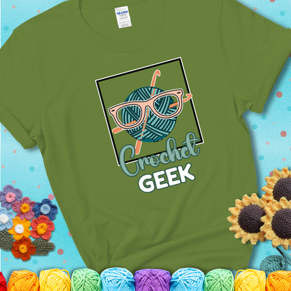 A teal yarn ball with crisscrossed hooks wears oversized nerdy glasses with the phrase “Crochet Geek” in bold, fun text below on a green t-shirt.