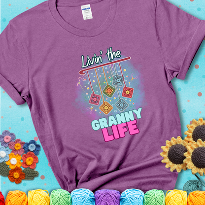 Light purple t-shirt shows colorful crochet granny squares hanging from a hook with text “Livin’ the Granny Life” in bold pink and blue font; a fun nod to classic crochet.