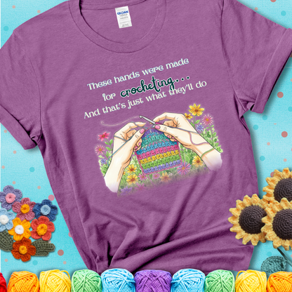 Heather Radiant Orchid t-shirt with an illustration of two hands crocheting a rainbow swatch, surrounded by colorful flowers and the phrase “These hands were made for crocheting… and that’s just what they’ll do.”