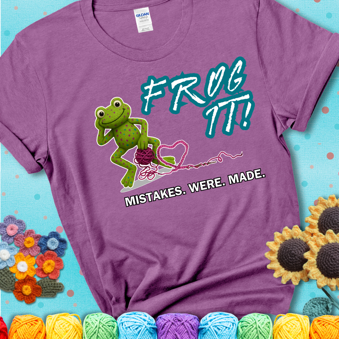 Green frog with yarn and heart-shaped loop. Text reads: “Frog It! Mistakes Were Made.” A playful crochet tee for anyone who's ever had to rip it back, showing a lavender tee with a teal sunflower background