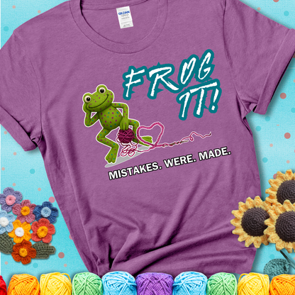 Green frog with yarn and heart-shaped loop. Text reads: “Frog It! Mistakes Were Made.” A playful crochet tee for anyone who's ever had to rip it back, showing a lavender tee with a teal sunflower background