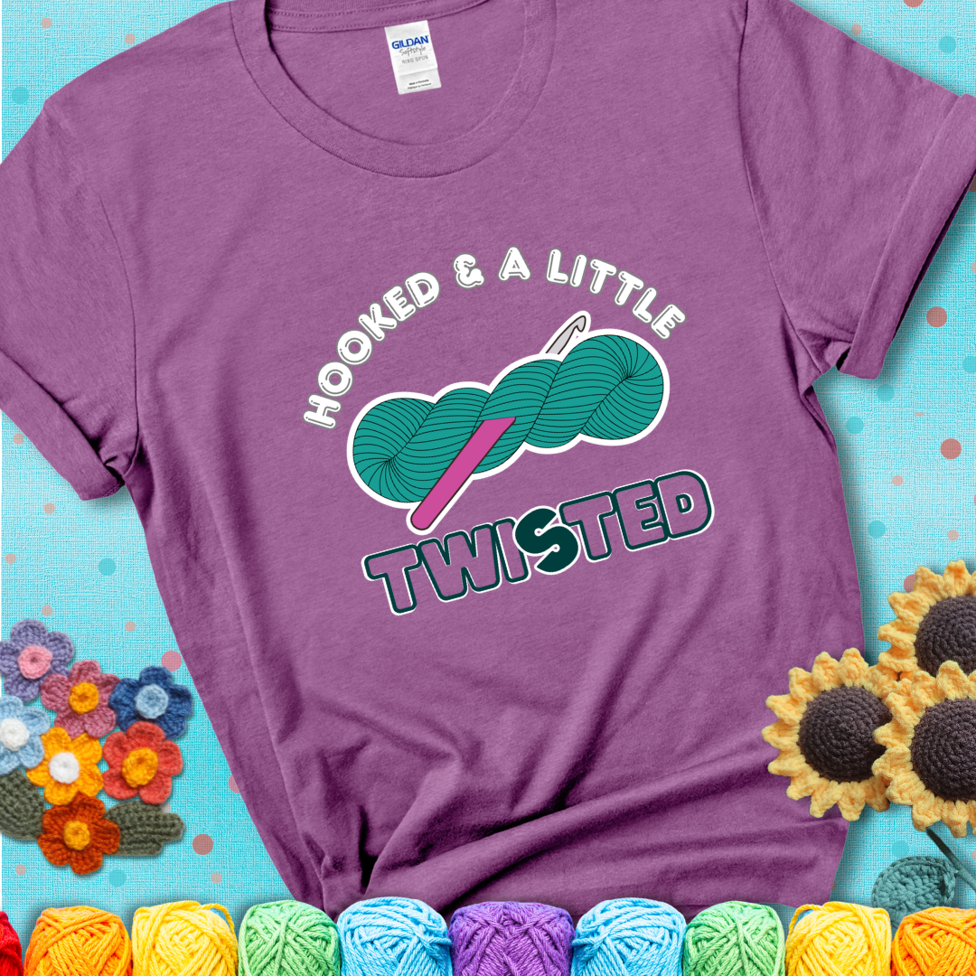 A teal skein of yarn with a hot pink crochet hook through it, captioned “Hooked & a Little Twisted” on a light purple t-shirt.