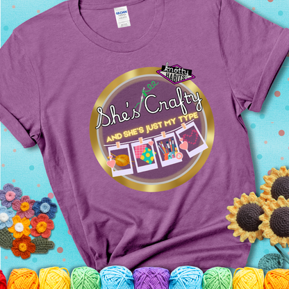 Text on light purple tee reads "She’s Crafty & She’s Just My Type" with yarn, fabric, painting, and cross-stitch icons hanging like Polaroids
