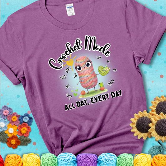 Light purple t-shirt with a kawaii-style rainbow yarn character surrounded by crochet stitch abbreviations and holding a yarn heart, with text “Crochet Mode All Day, Every Day.”