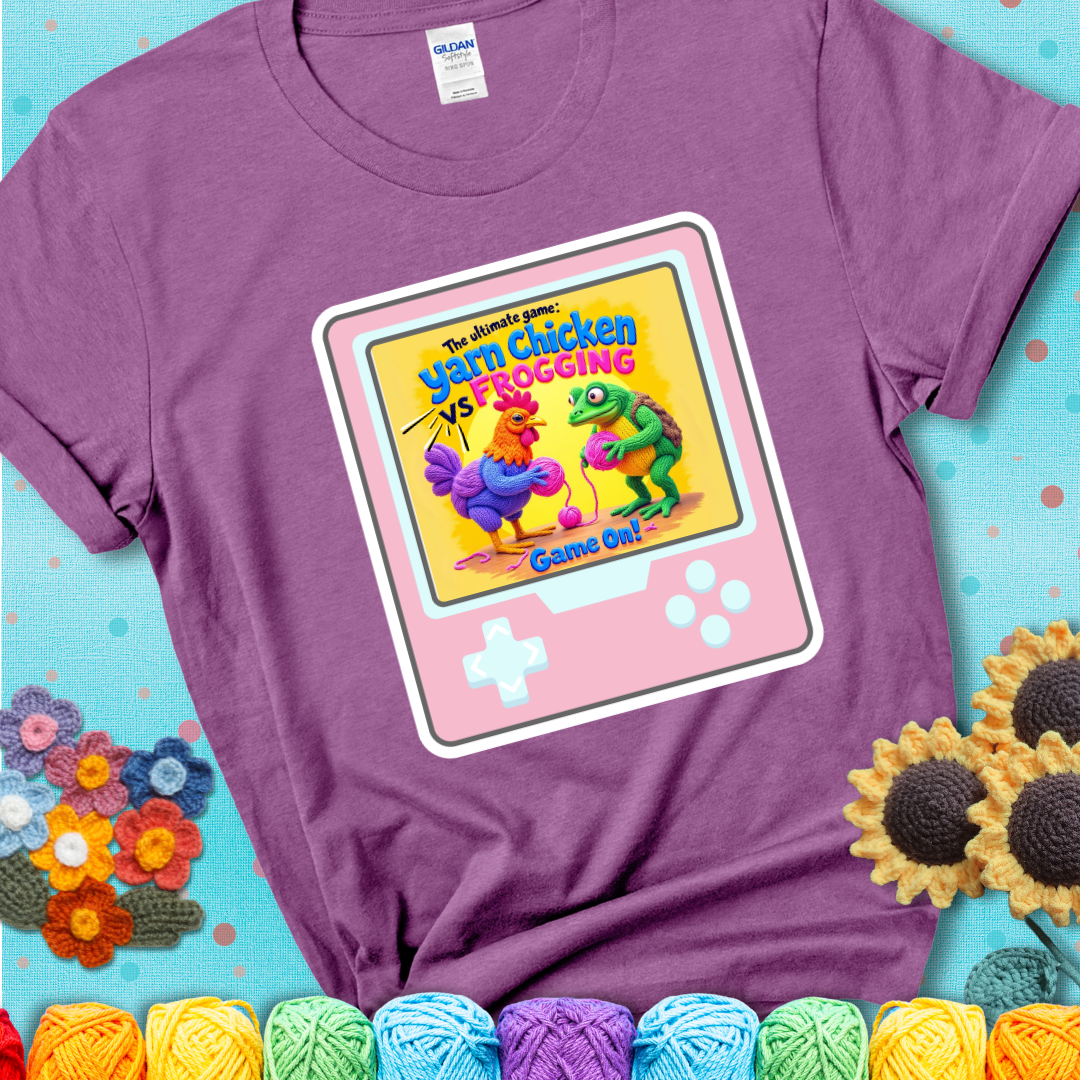 A light purple t-shirt featuring a cartoon yarn chicken and frog face off with text “Yarn Chicken vs Frogging: Game On!” on a retro, handheld video game meets crochet humor battle design.