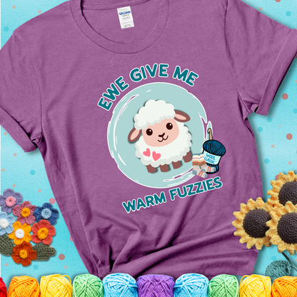 Light purple t-shirt showing a cartoon sheep with pink hearts, wool socks, and yarn labeled “Wool Classic.” Text reads “Ewe Give Me Warm Fuzzies.”