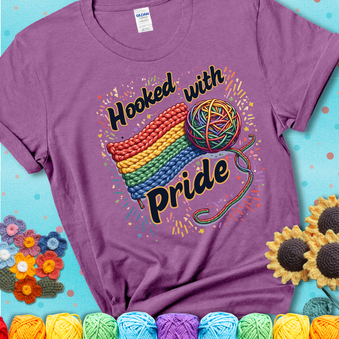 A rainbow-striped crochet swatch paired with a multicolor yarn ball and bold “Hooked With Pride” lettering. Confetti surrounds the vibrant scene showing a lavender tee with a teal sunflower background