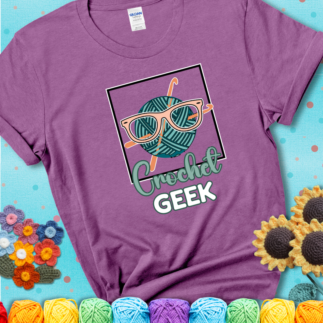 A teal yarn ball with crisscrossed hooks wears oversized nerdy glasses with the phrase “Crochet Geek” in bold, fun text below on a light purple t-shirt.