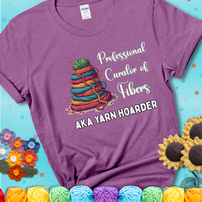 A light purple t-shirt showing a pyramid of rainbow yarn topped with green ball; text reads “Professional Curator of Fibers AKA Yarn Hoarder”, a playful shirt for serious stashers.