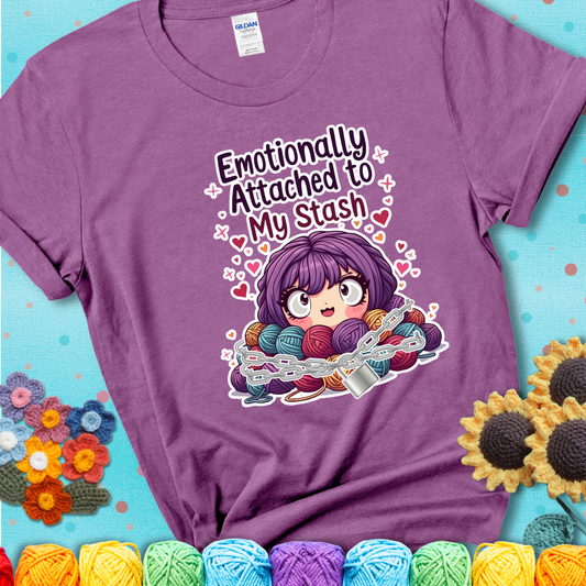 Adorable character with wide eyes hugging a yarn stash wrapped in chains and a lock, with the text “Emotionally Attached to My Stash”  on a lavender tee.