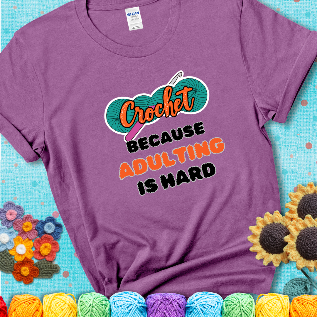 Light purple t-shirt featuring a bold design with the phrase “Crochet Because Adulting Is Hard” featuring a green skein of yarn with a pink crochet hook. The word “Crochet” is in a script font, while “Adulting is hard” is in chunky textured letters.