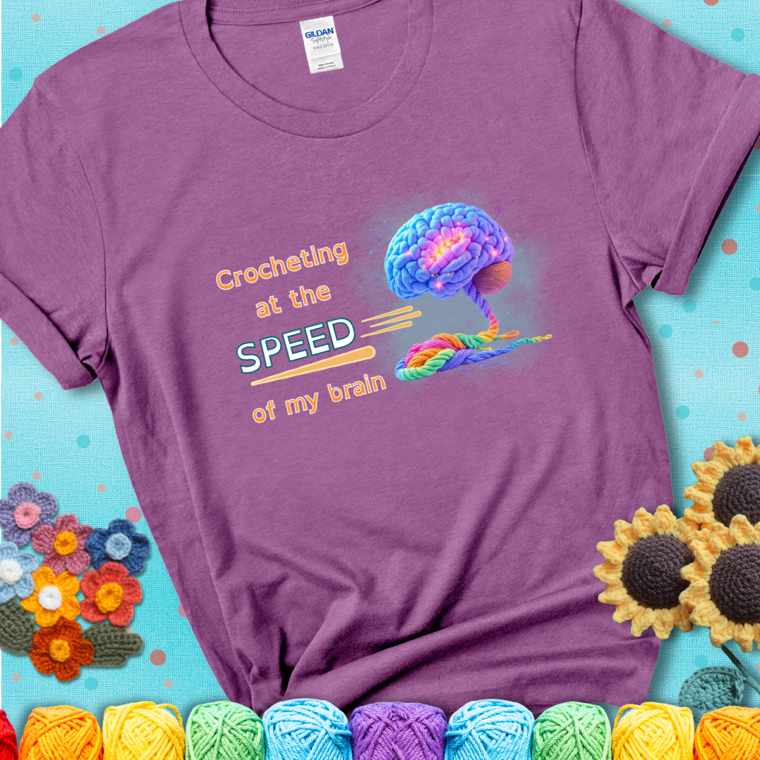 A glowing rainbow brain unravels into colorful yarn with the phrase “Crocheting at the speed of my brain” on a light purple t-shirt. 