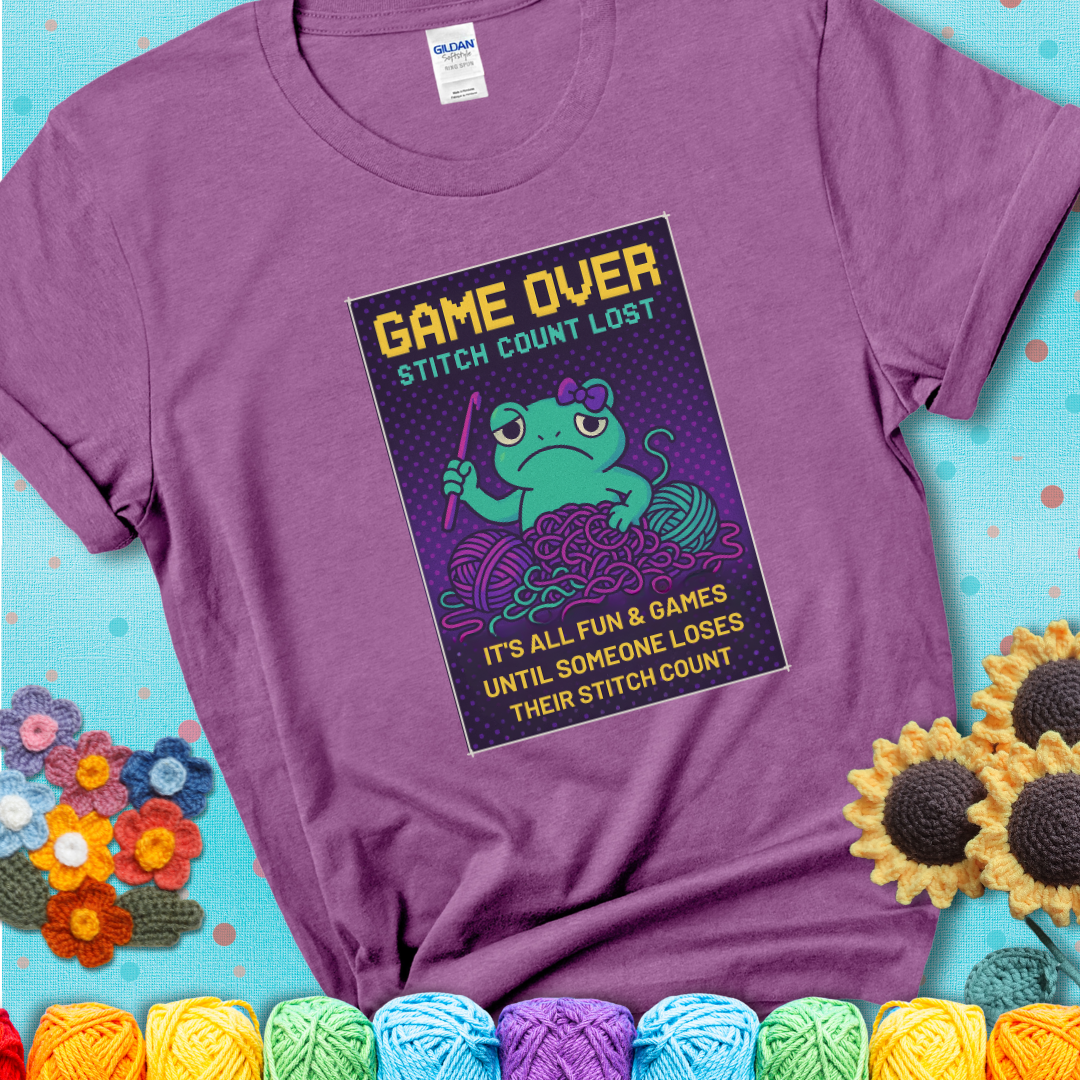 Light purple t-shirt shows a pixel-style design of a grumpy yarn frog holding a hook, surrounded by yarn chaos, with the text “Game Over. Stitch Count Lost.”