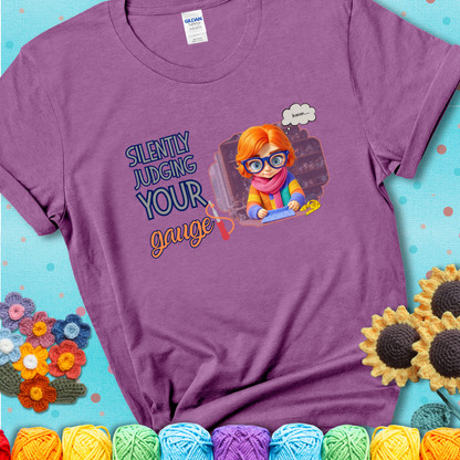Heather Radiant Orchid t-shirt with a serious-looking crocheter in rainbow clothing and big glasses, holding a tape measure under the phrase “Silently Judging Your Gauge.”