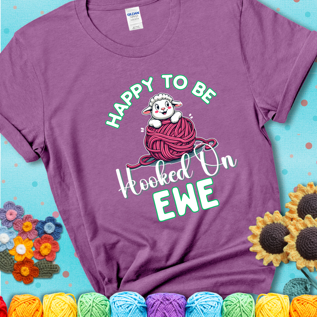 Light purple t-shirt with smiling cartoon sheep peeks from a pink yarn ball, surrounded by green text “Happy to Be Hooked on Ewe”, a playful and affectionate sheep pun for crocheters.