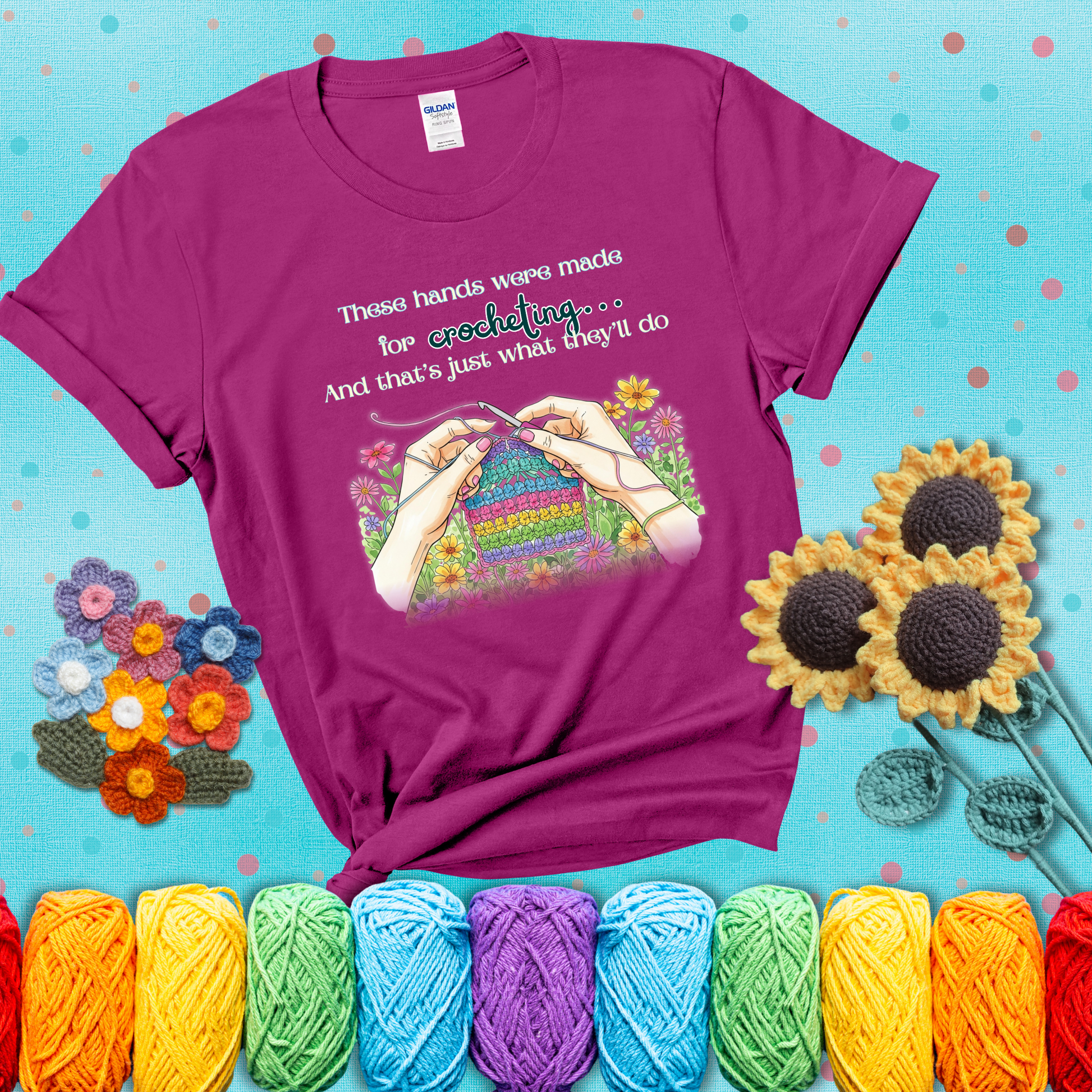 Illustration of two hands crocheting a rainbow swatch, surrounded by vibrant flowers with the phrase “These hands were made for crocheting… and that’s just what they’ll do,” showing a magenta tee with a teal sunflower background