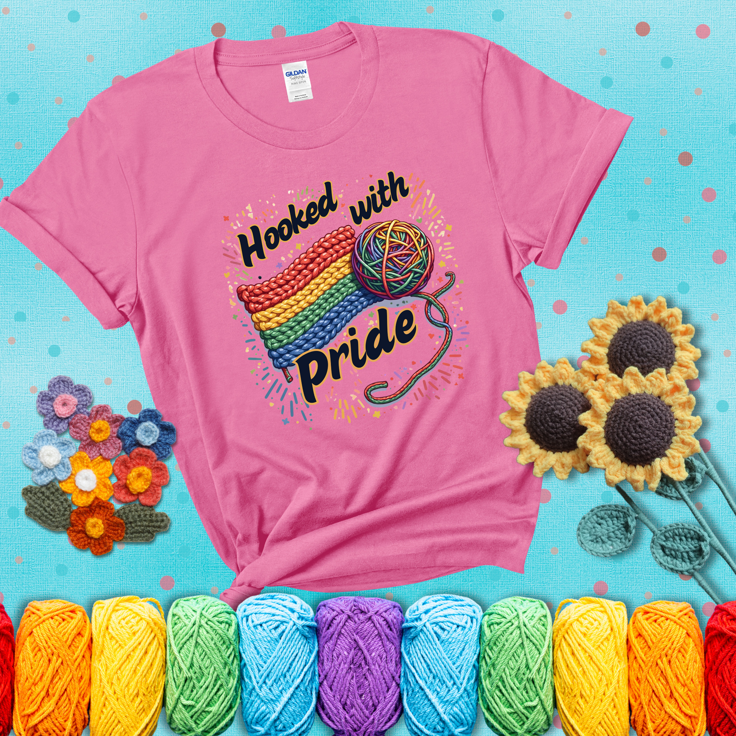 A rainbow-striped crochet swatch paired with a multicolor yarn ball and bold “Hooked With Pride” lettering. Confetti surrounds the vibrant scene showing a pink tee with a teal sunflower background