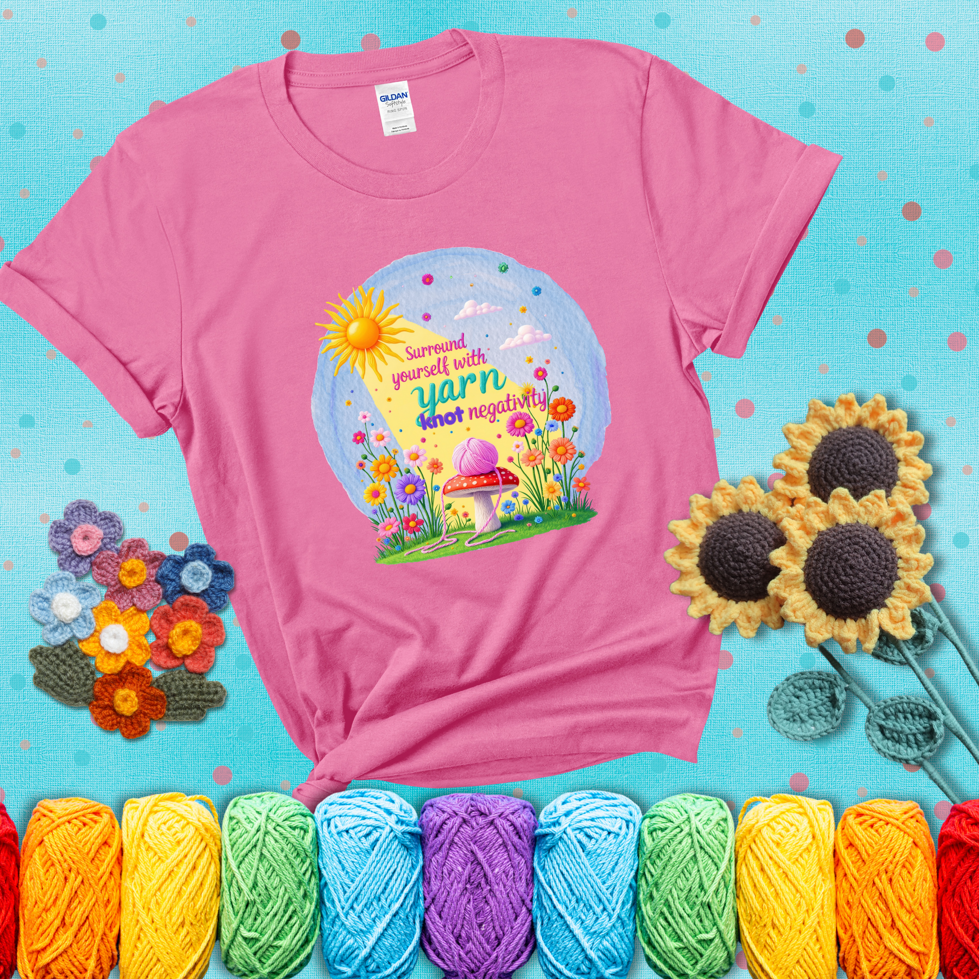 A happy spring garden scene with a sunbeam, flowers, and a pink yarn ball on a mushroom, with the quote "Surround yourself with yarn, knot negativity," showing a pink tee with a teal sunflower background