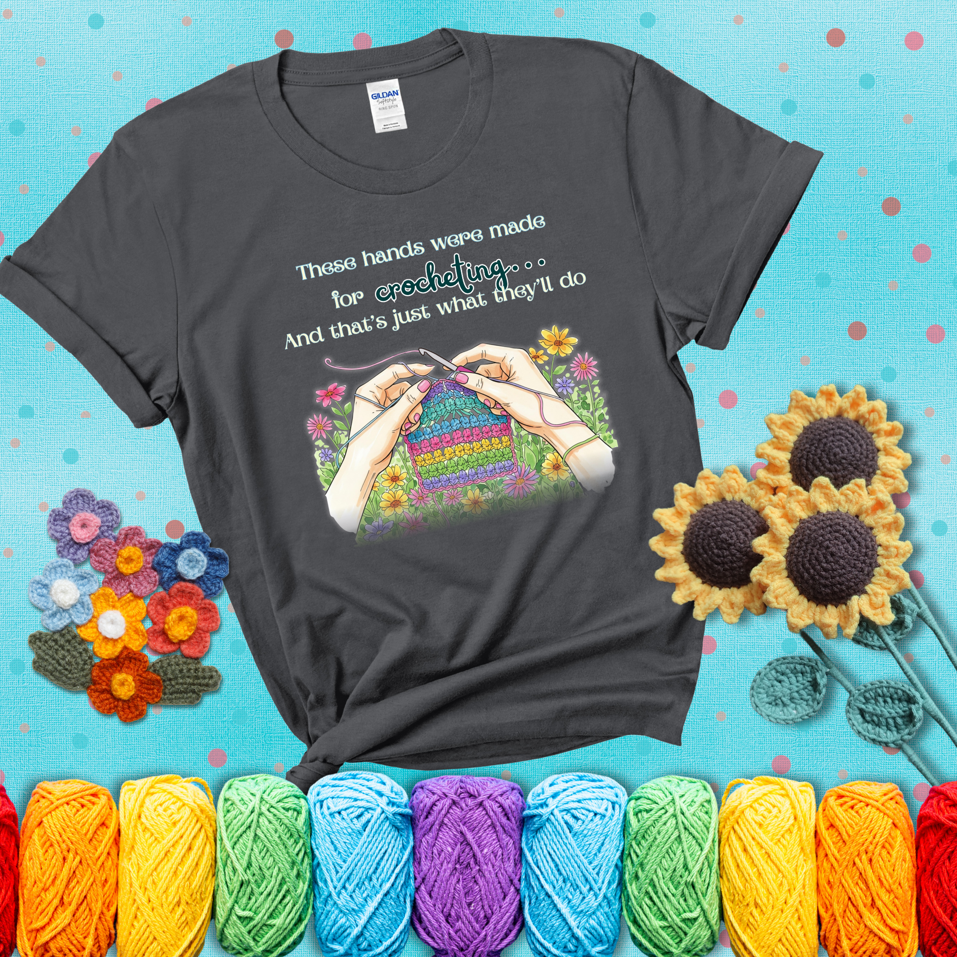 Illustration of two hands crocheting a rainbow swatch, surrounded by vibrant flowers with the phrase “These hands were made for crocheting… and that’s just what they’ll do,” showing a dark grey tee with a teal sunflower background