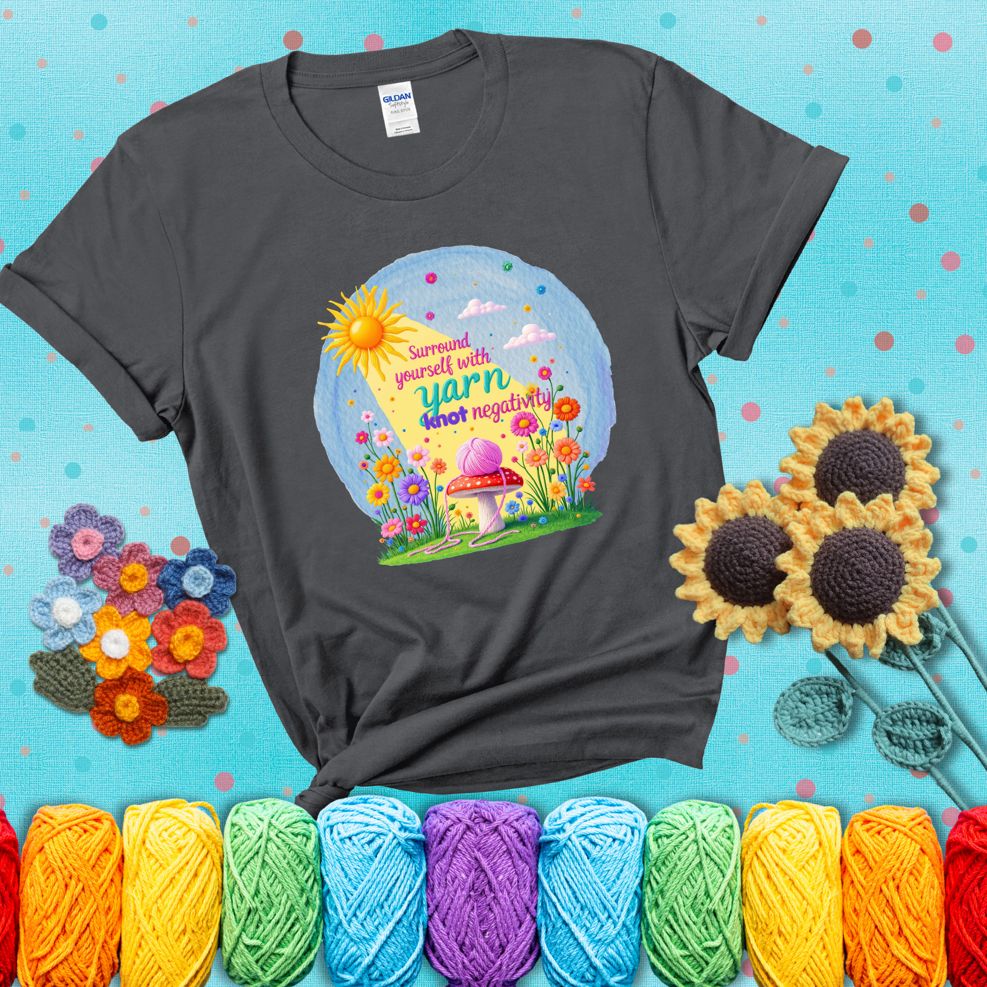 A happy spring garden scene with a sunbeam, flowers, and a pink yarn ball on a mushroom, with the quote "Surround yourself with yarn, knot negativity," showing a dark grey tee with a teal sunflower background