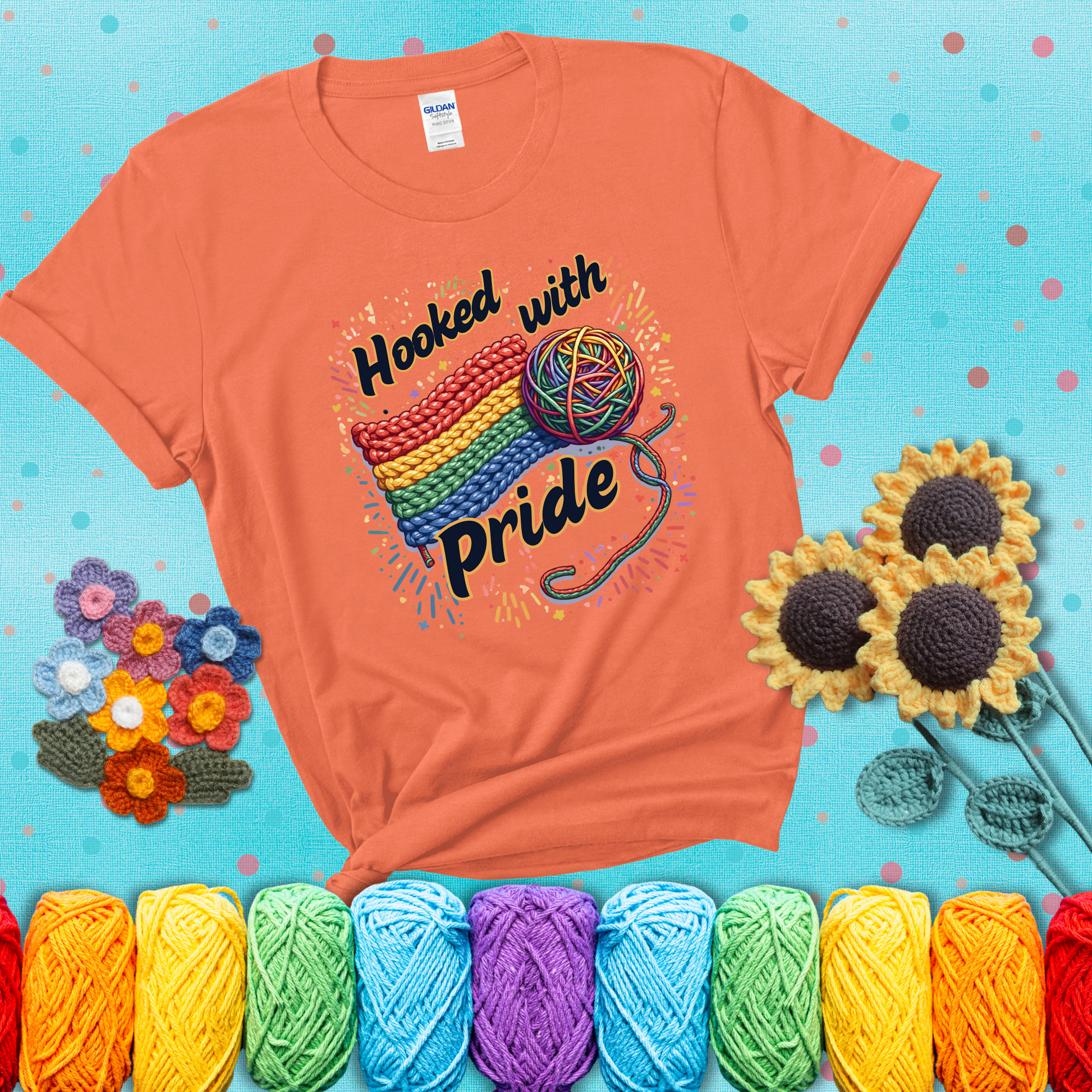 A rainbow-striped crochet swatch paired with a multicolor yarn ball and bold “Hooked With Pride” lettering. Confetti surrounds the vibrant scene showing a light orange tee with a teal sunflower background