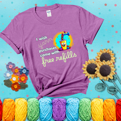 A bright yellow cup with a smiling face is being filled with colorful yarn balls from a soda machine, with the phrase “I wish yarn purchases came with free refills,” showing a light purple tee with a teal sunflower background