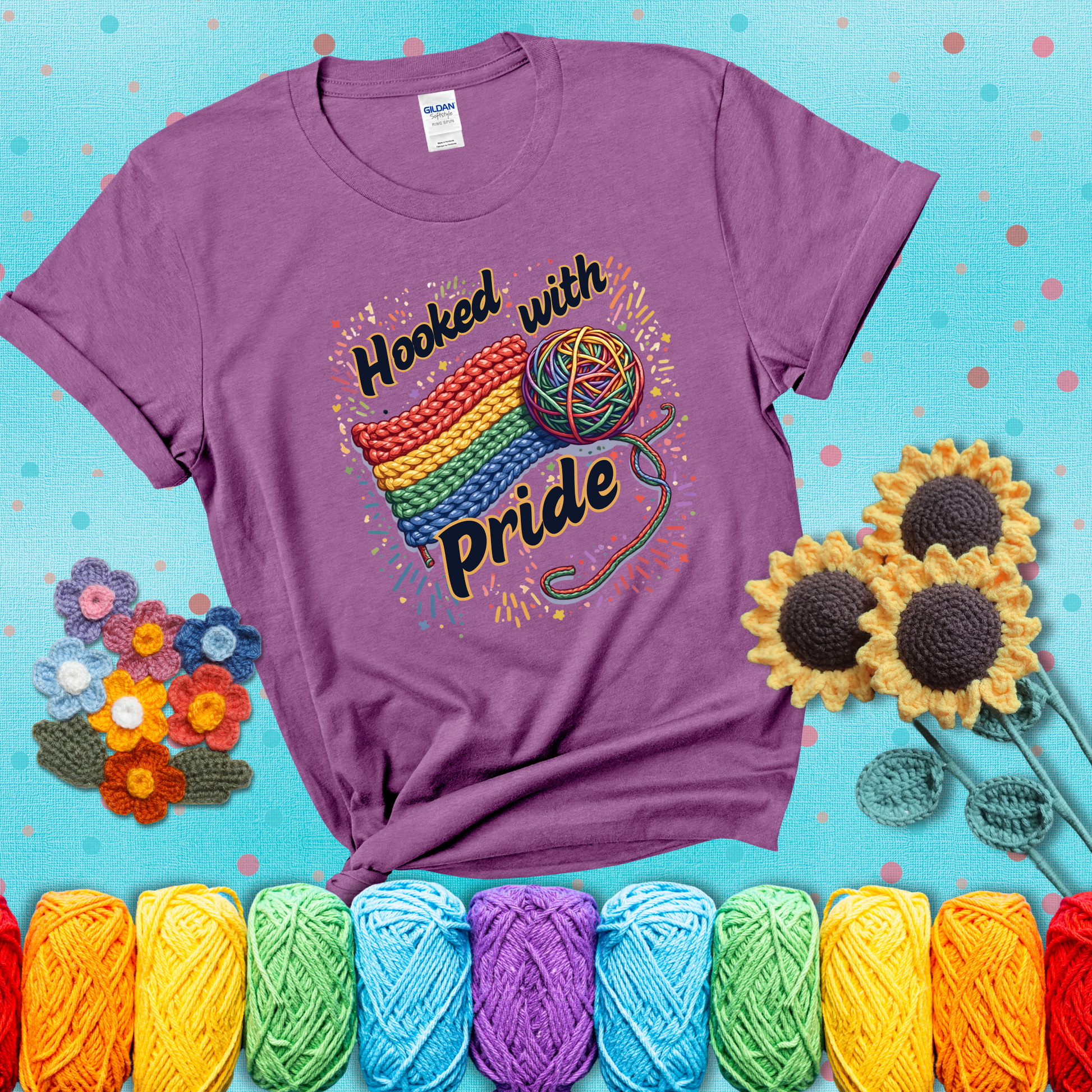 A rainbow-striped crochet swatch paired with a multicolor yarn ball and bold “Hooked With Pride” lettering. Confetti surrounds the vibrant scene showing a light purple tee with a teal sunflower background