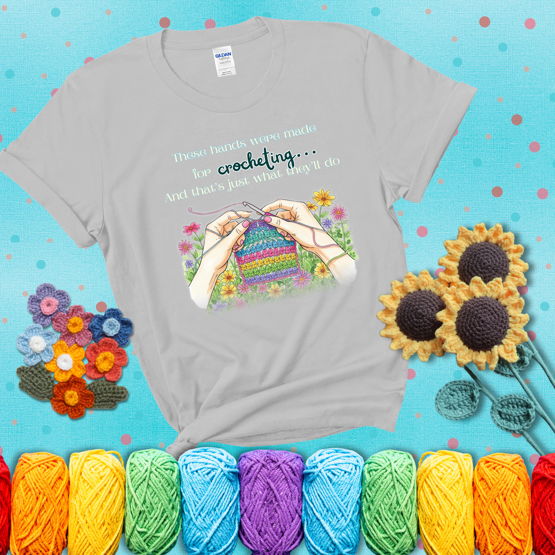 Illustration of two hands crocheting a rainbow swatch, surrounded by vibrant flowers with the phrase “These hands were made for crocheting… and that’s just what they’ll do,” showing a light grey tee with a teal sunflower background