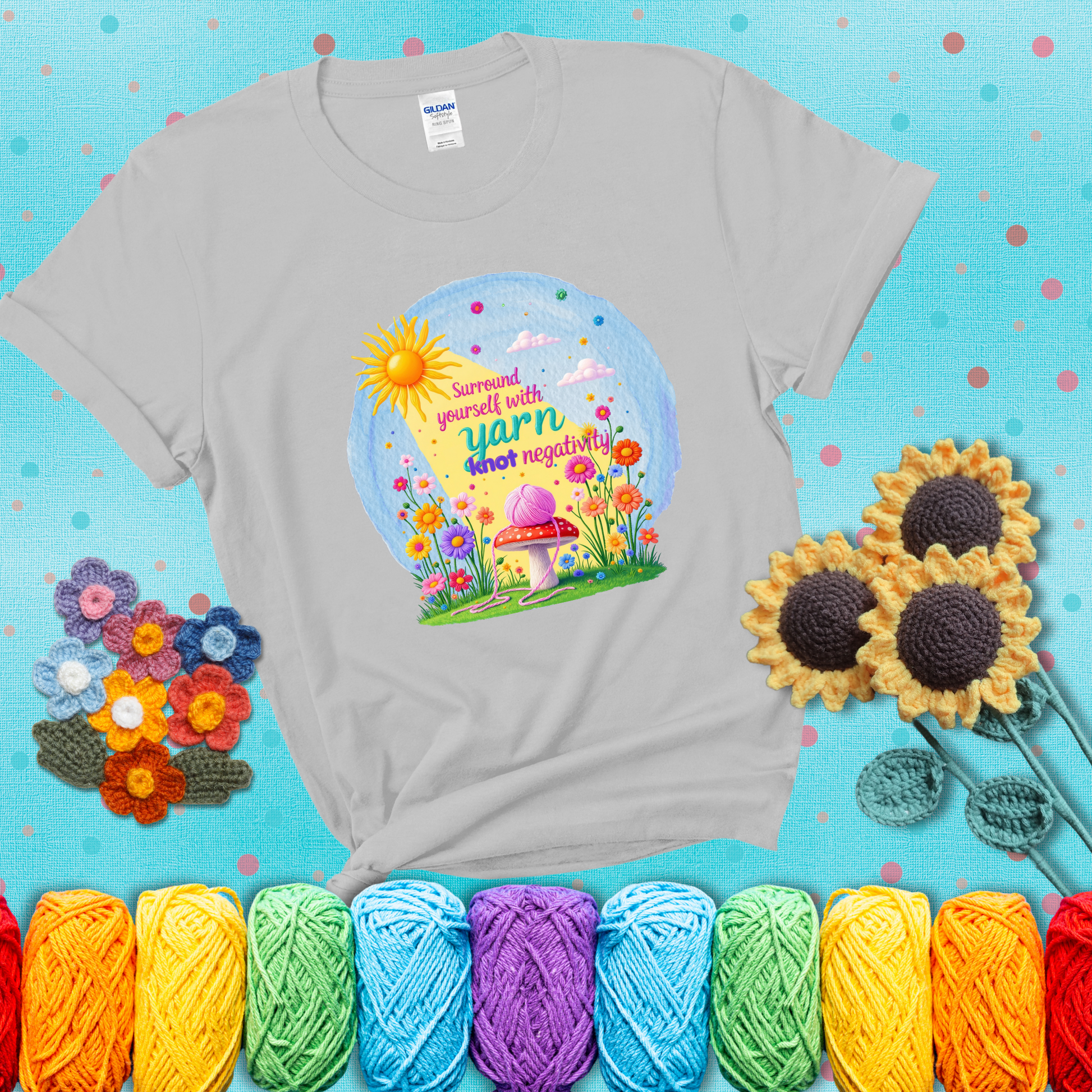 A happy spring garden scene with a sunbeam, flowers, and a pink yarn ball on a mushroom, with the quote "Surround yourself with yarn, knot negativity," showing a light grey tee with a teal sunflower background