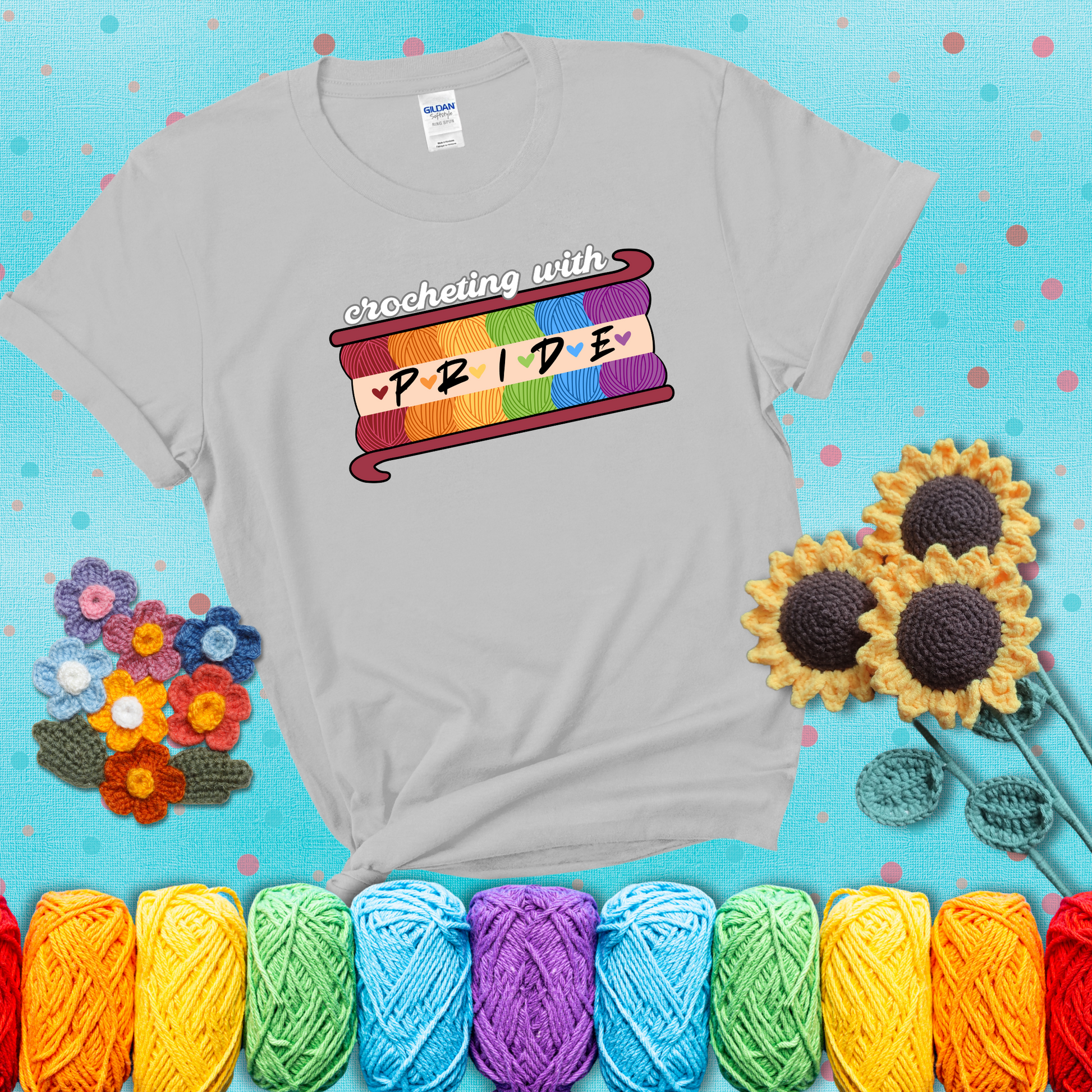Yarn skeins in rainbow colors stacked in a hook shape. Text reads “Crocheting With Pride” showing a light grey tee with a teal sunflower background