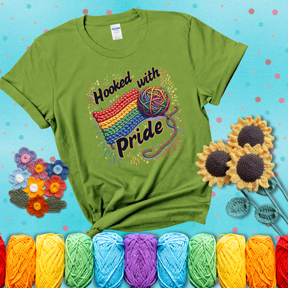 A rainbow-striped crochet swatch paired with a multicolor yarn ball and bold “Hooked With Pride” lettering. Confetti surrounds the vibrant scene showing a bright green tee with a teal sunflower background